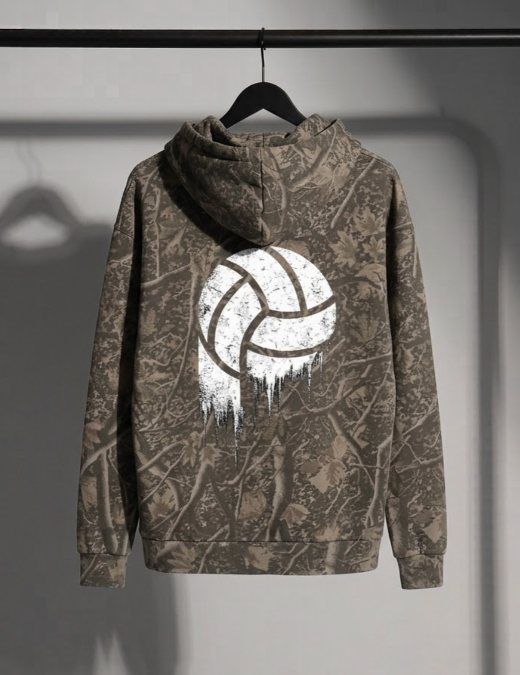 A camouflage hoodie hanging on a black hanger against a gray wall. The hoodie features a brown and gray woodland camo pattern with a large white distressed faded volleyball graphic printed on the back.