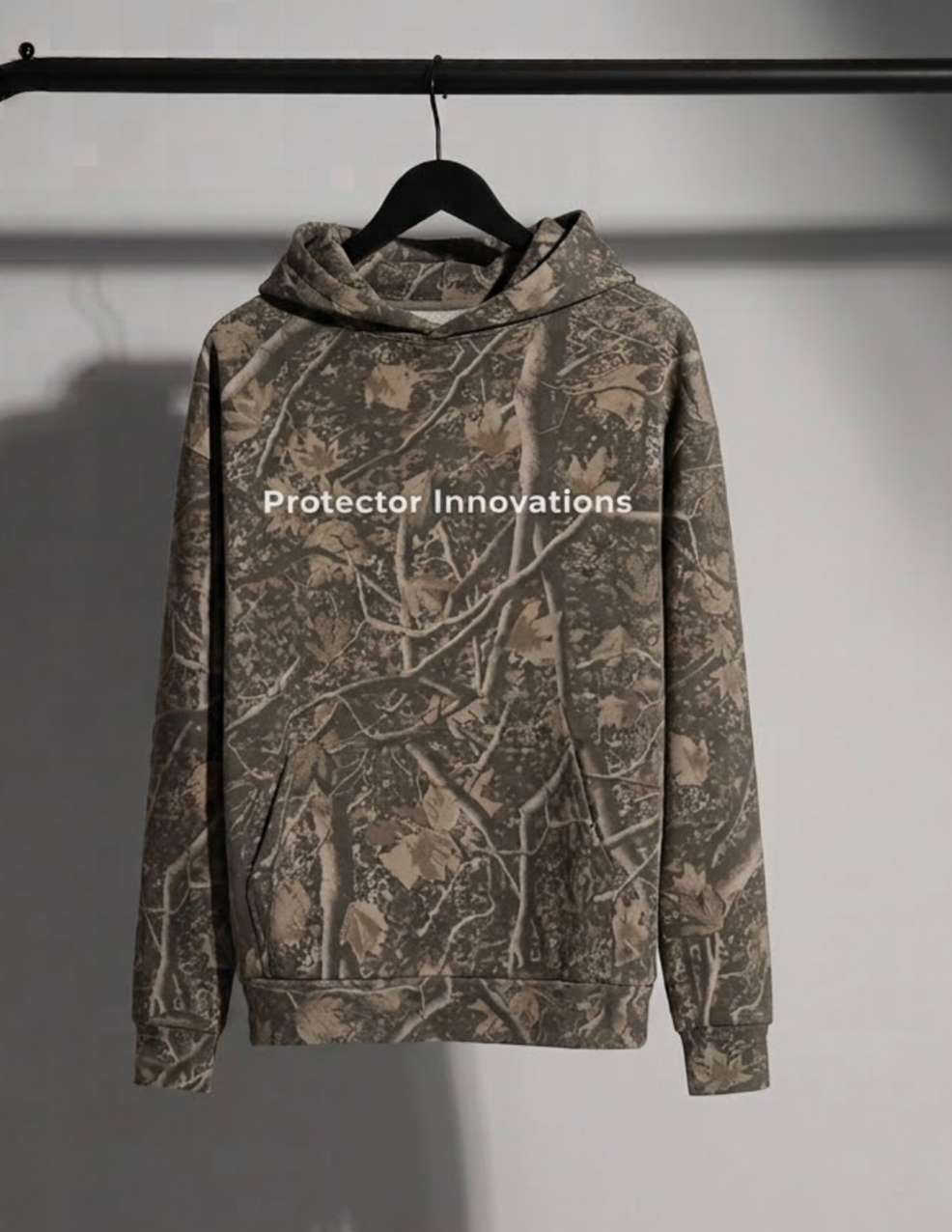 A camouflage hoodie with a brown and gray woodland branch pattern, hanging on a black hanger from a black clothing rack against a gray wall. The brand name 'Protector Innovations' is printed in white text across the chest.