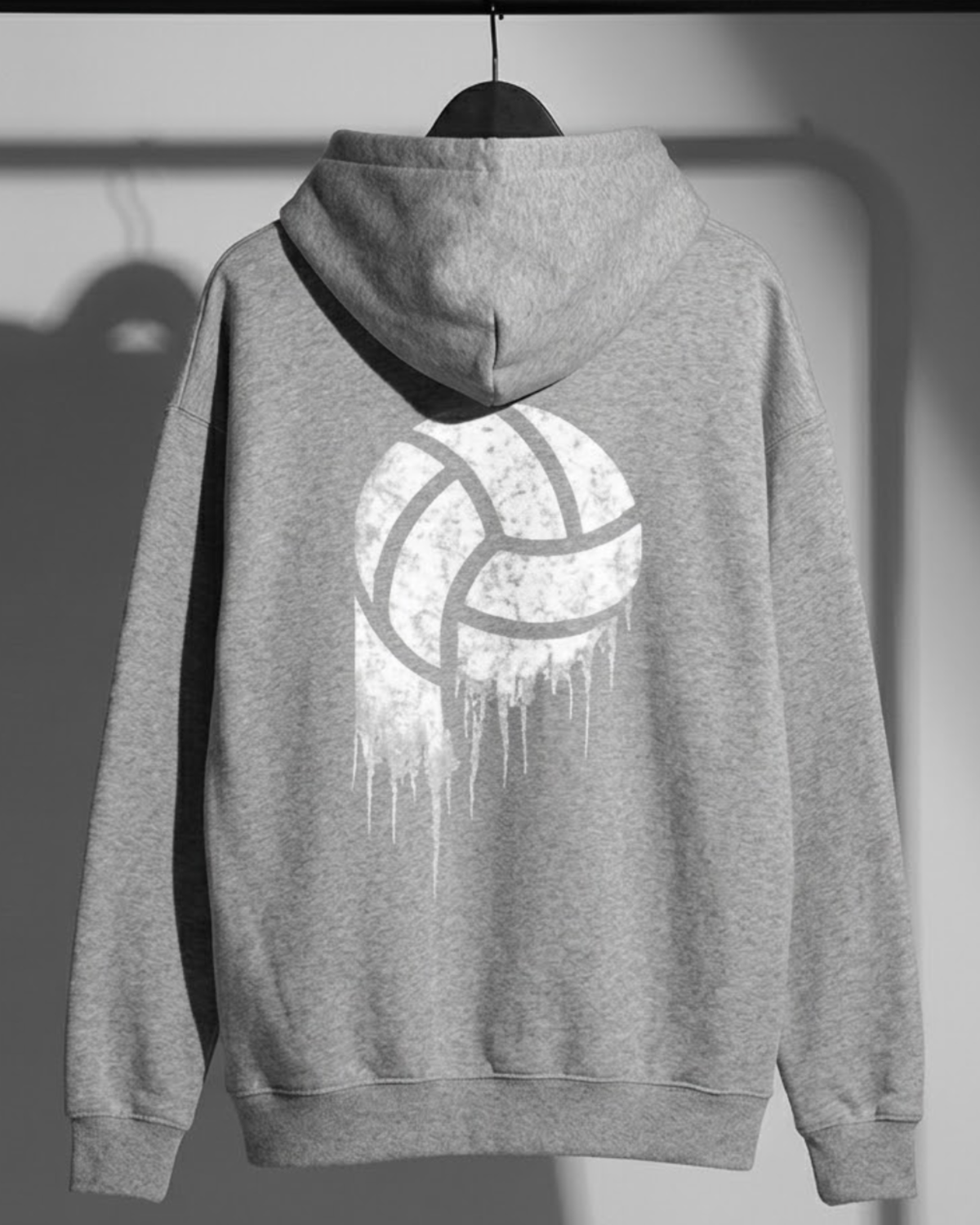 A heather gray pullover hoodie hanging on a black hanger against a light gray wall. The back of the hoodie features a large distressed white volleyball graphic with a dripping paint effect.