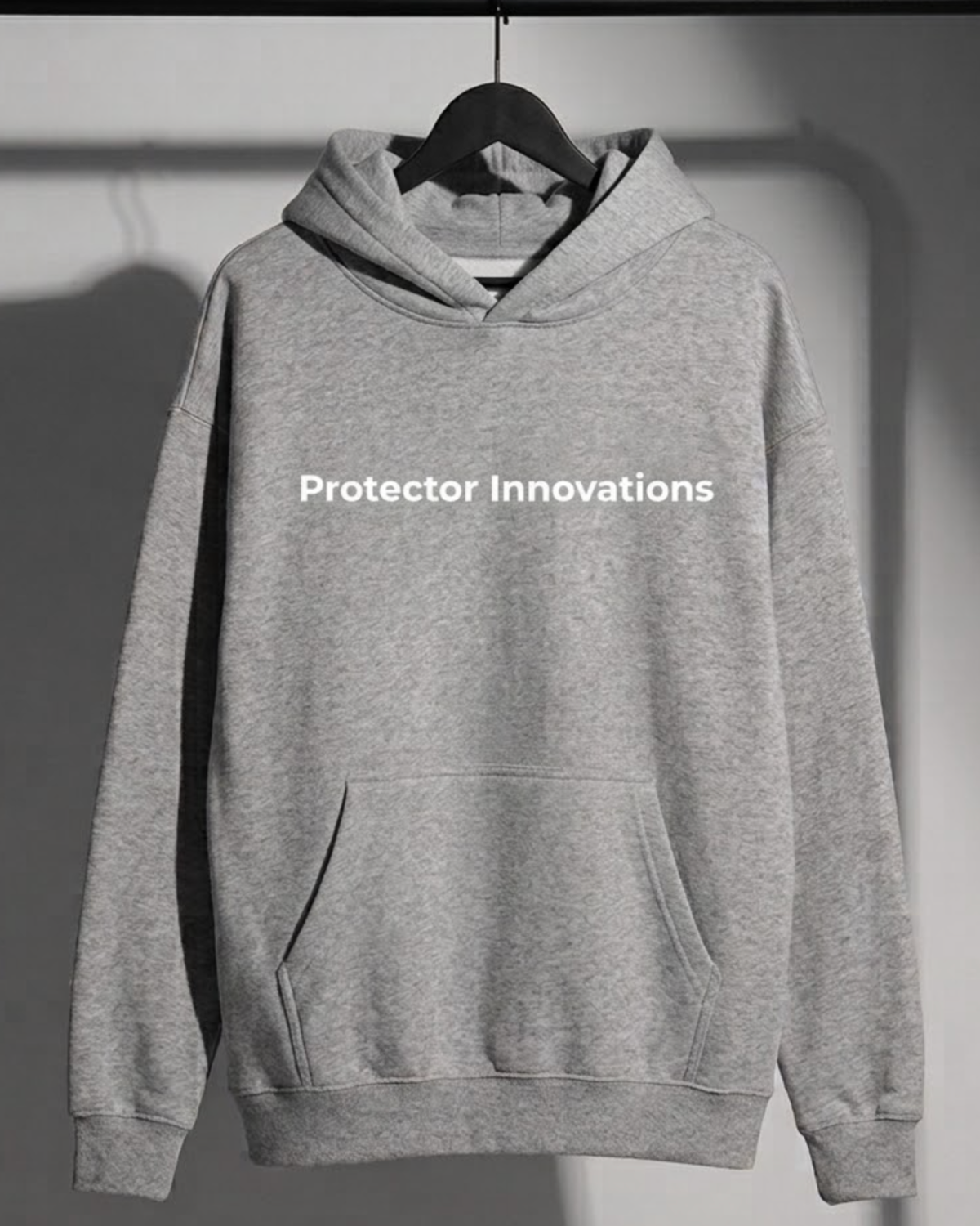 A light gray heather pullover hoodie hanging on a black hanger against a dark gray wall. The hoodie features the text 'Protector Innovations' printed in white across the chest.